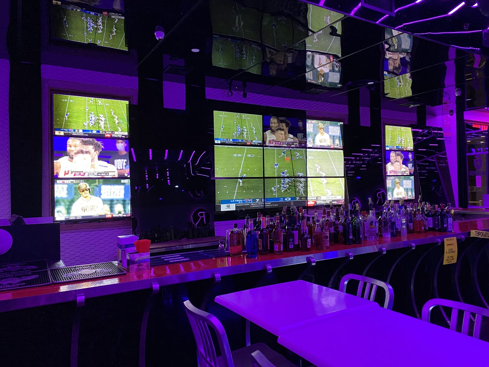 A wall of screens showing multiple games above a bar under violet lighting