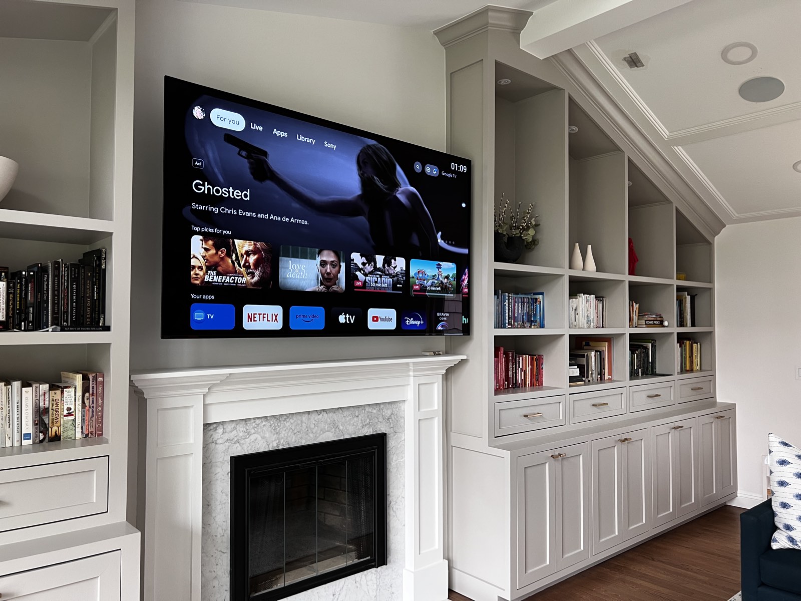 A large display mounted above a marble fireplace between built-in shelving, its home screen up